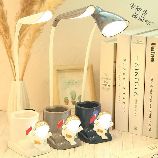 Desk lamp with decorative figures and books on a wooden surface. Astronaut LED Desk Lamp with Penholder - Madina Gift